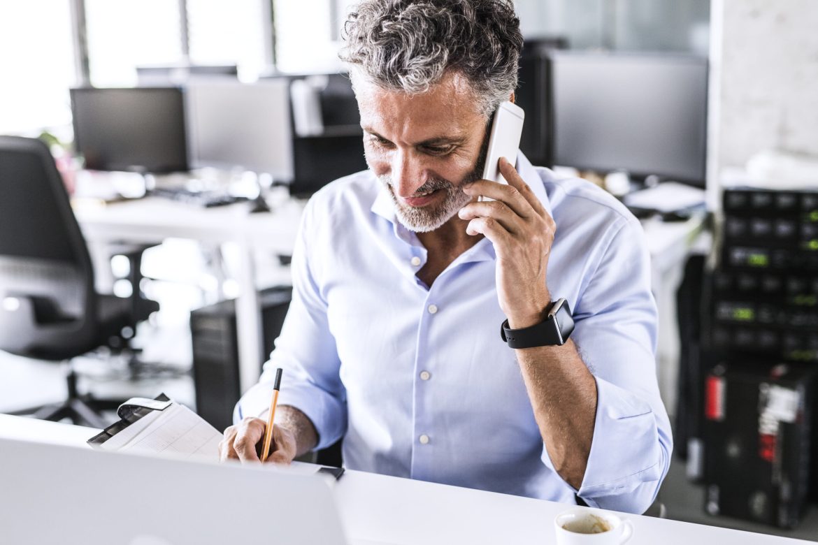 Smiling mature businessman sitting at desk in office talking on smartphone